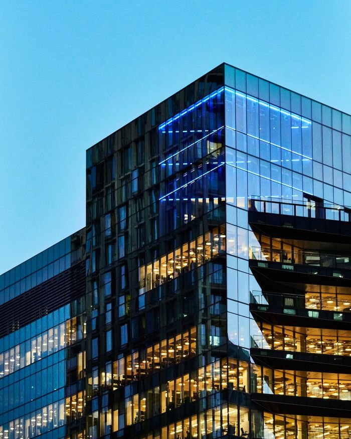 Mengenal Pentingnya Perizinan Bangunan Sebelum Proyek Berjalan A sleek glass office building with illuminated interior visible against a twilight sky.